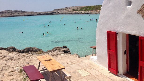 A beachside scene with a white building, a wooden table, and red shutters. - Hotel Romantic Los 5 Sentidos (Ciutadella de Menorca)