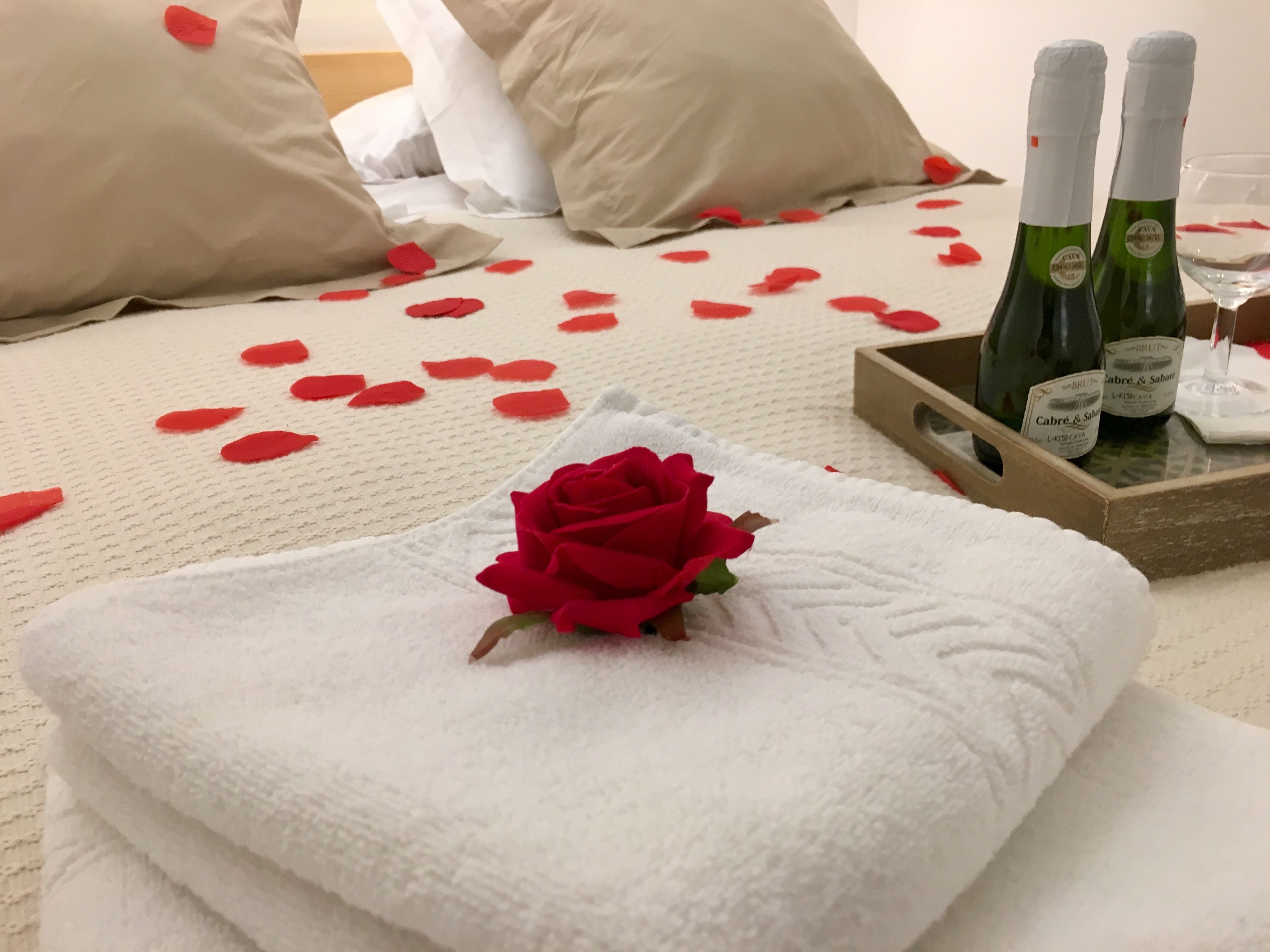 A neatly made bed with rose petals, a rose, and two bottles of champagne on a tray.