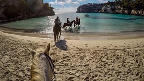 A beach with clear turquoise water, a horse in the foreground, and people riding horses in the background. - Hotel Romantic Los 5 Sentidos (Ciutadella de Menorca)