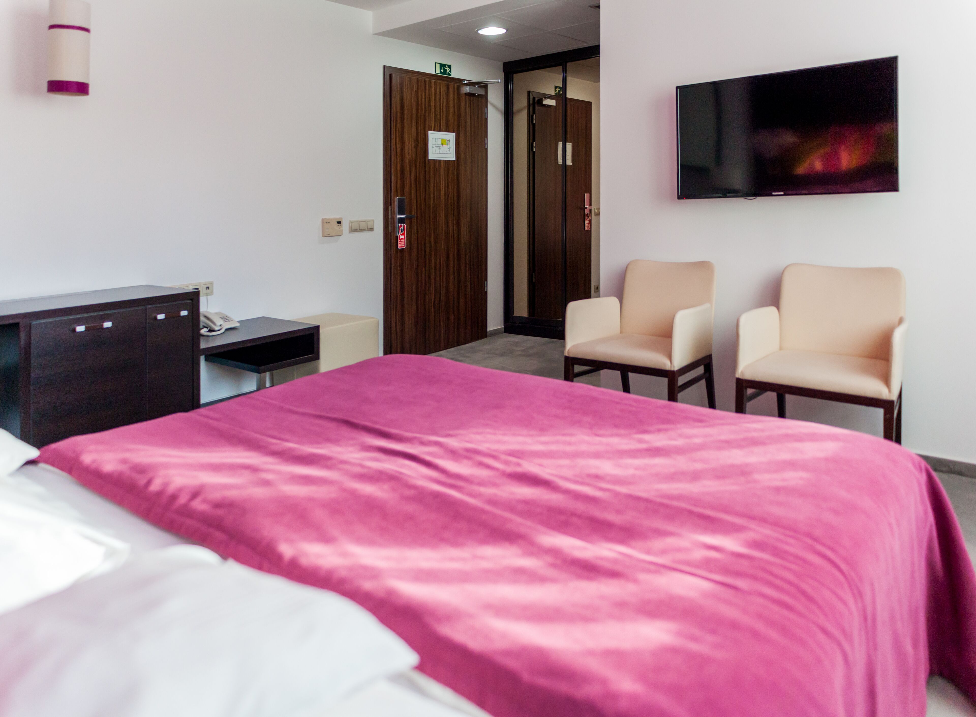 Habitación de hotel con cama, dos sillas, escritorio con teléfono, televisión y puerta con señal de salida.