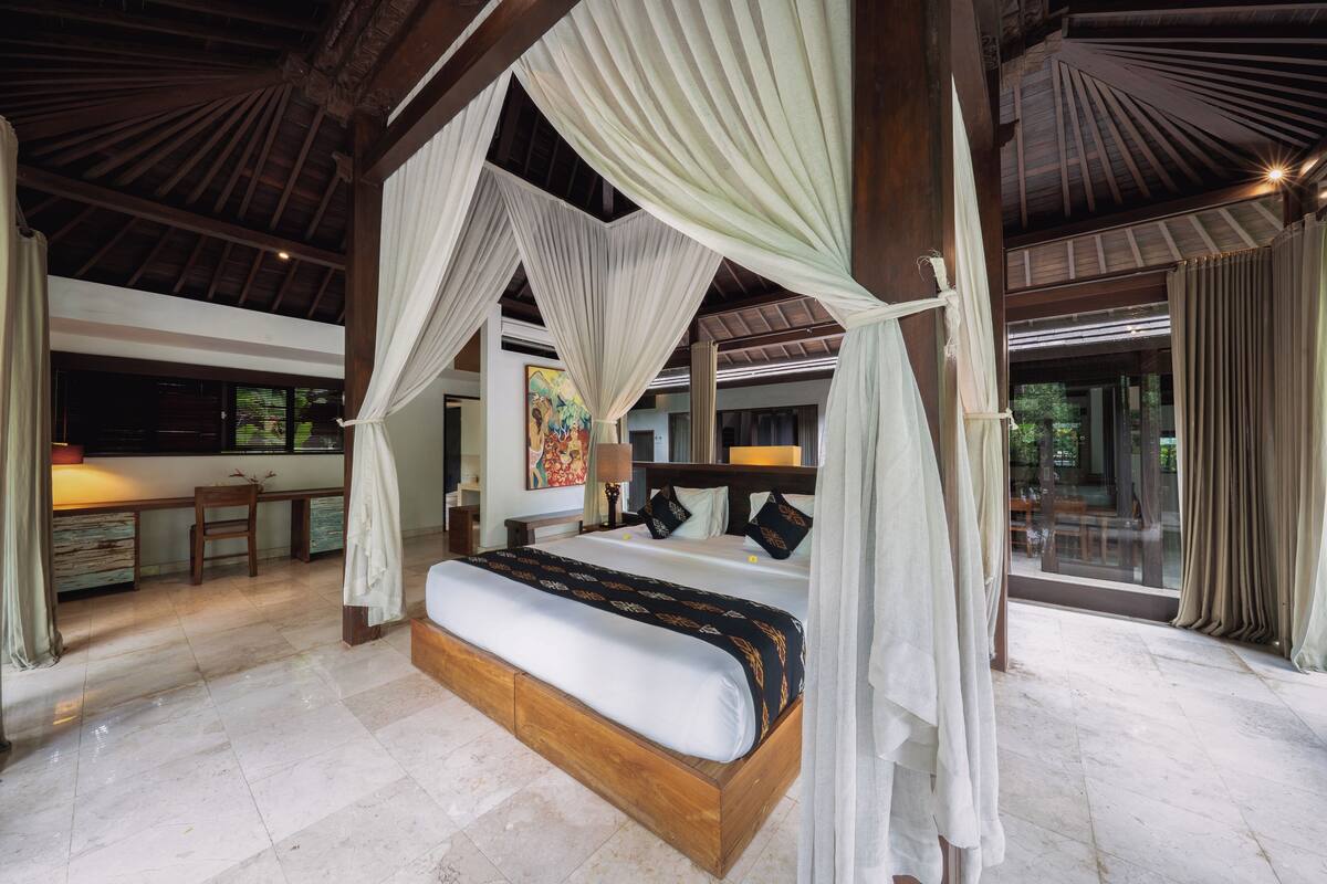 A spacious bedroom with a large bed, a wooden headboard, and a canopy. There is a desk and chair in the corner, and a painting on the wall.