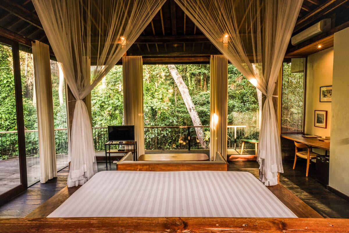 A spacious bedroom with a large bed, wooden flooring, and a balcony with a view of lush greenery.