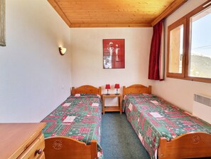 Een kamer met twee houten bedden, een houten dressoir, een wandlamp, een raam met rode gordijnen en een schilderij aan de muur.