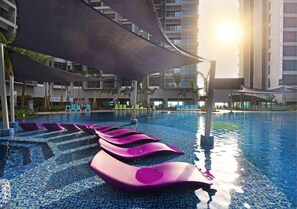 A swimming pool with floating exercise mats, a shaded area, and high-rise buildings in the background.