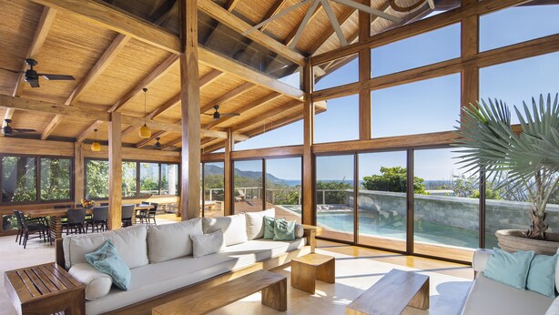 A spacious indoor area with a large wooden ceiling, a white sofa with cushions, wooden benches, and a view of a swimming pool through large windows. - Luxury Villa Melody- Santa Teresa beach  (Playa Santa Teresa)
