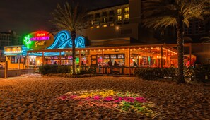 A brightly lit restaurant with a neon sign, outdoor seating, and palm trees.