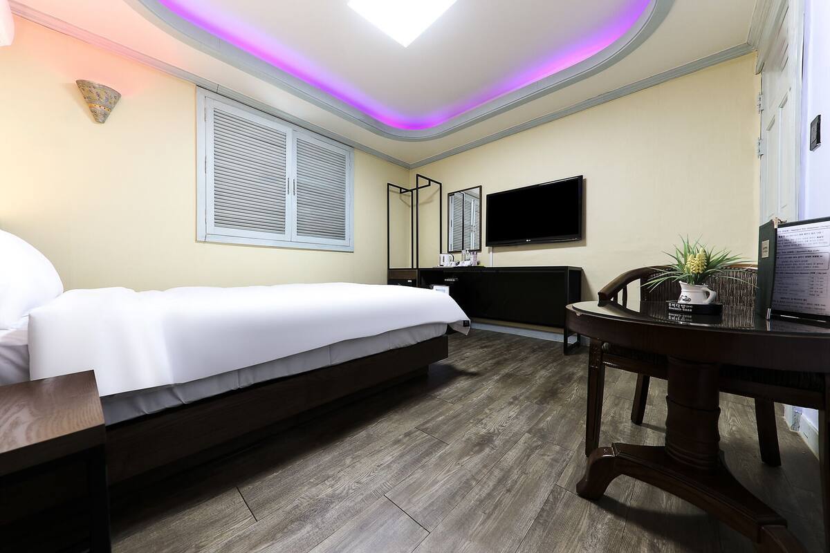 A hotel room with a bed, a desk, a chair, a TV, and a window with shutters.
