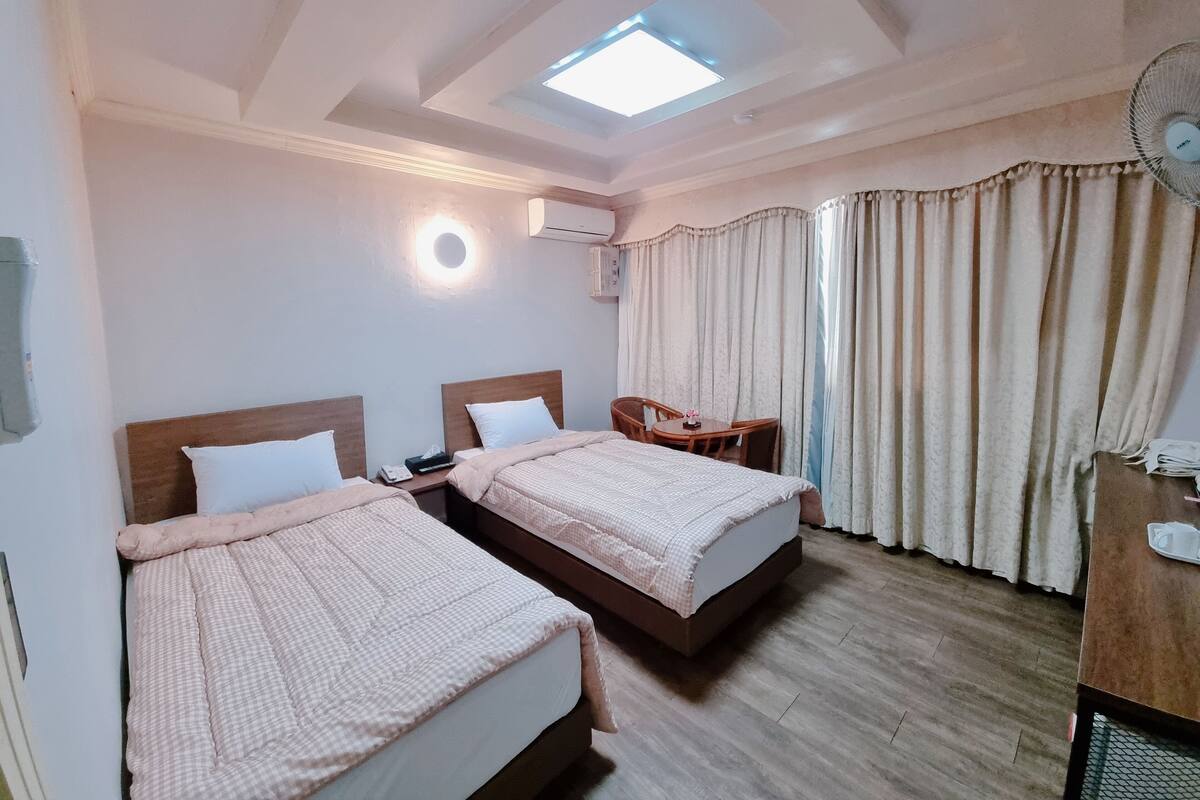 A hotel room with two beds, a small table, a chair, a ceiling fan, and a skylight.