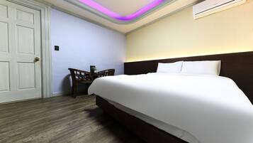 A hotel room with a large bed, a desk with a chair, a door, and an air conditioning unit on the wall.