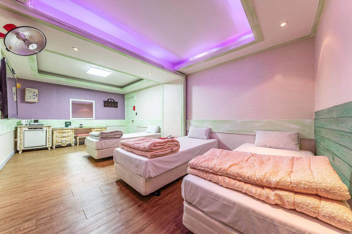 A hotel room with two beds, a desk, a chair, a small kitchenette, and a ceiling fan.