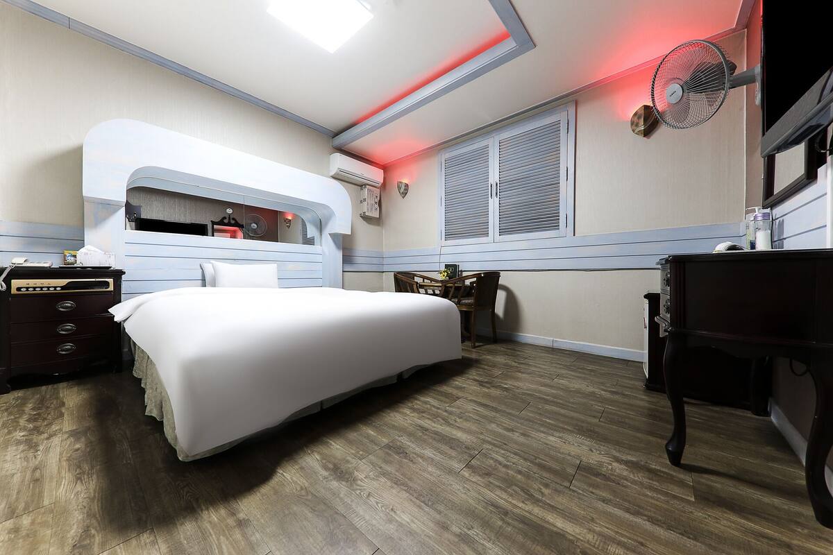 A hotel room with a bed, a desk, a chair, a dresser, a mirror, an air conditioning unit, and a window with blinds.