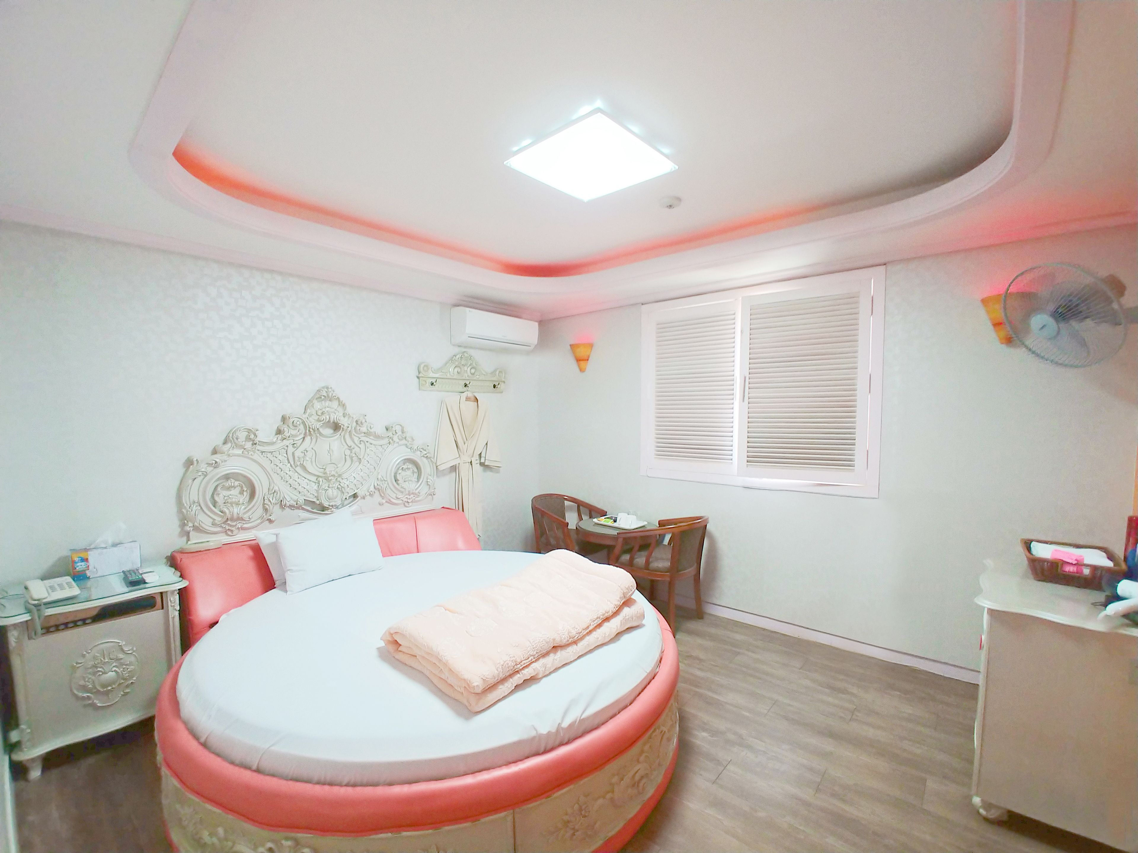 A bedroom with a round bed, a bedside table, a chair, a small table, and a window with blinds.