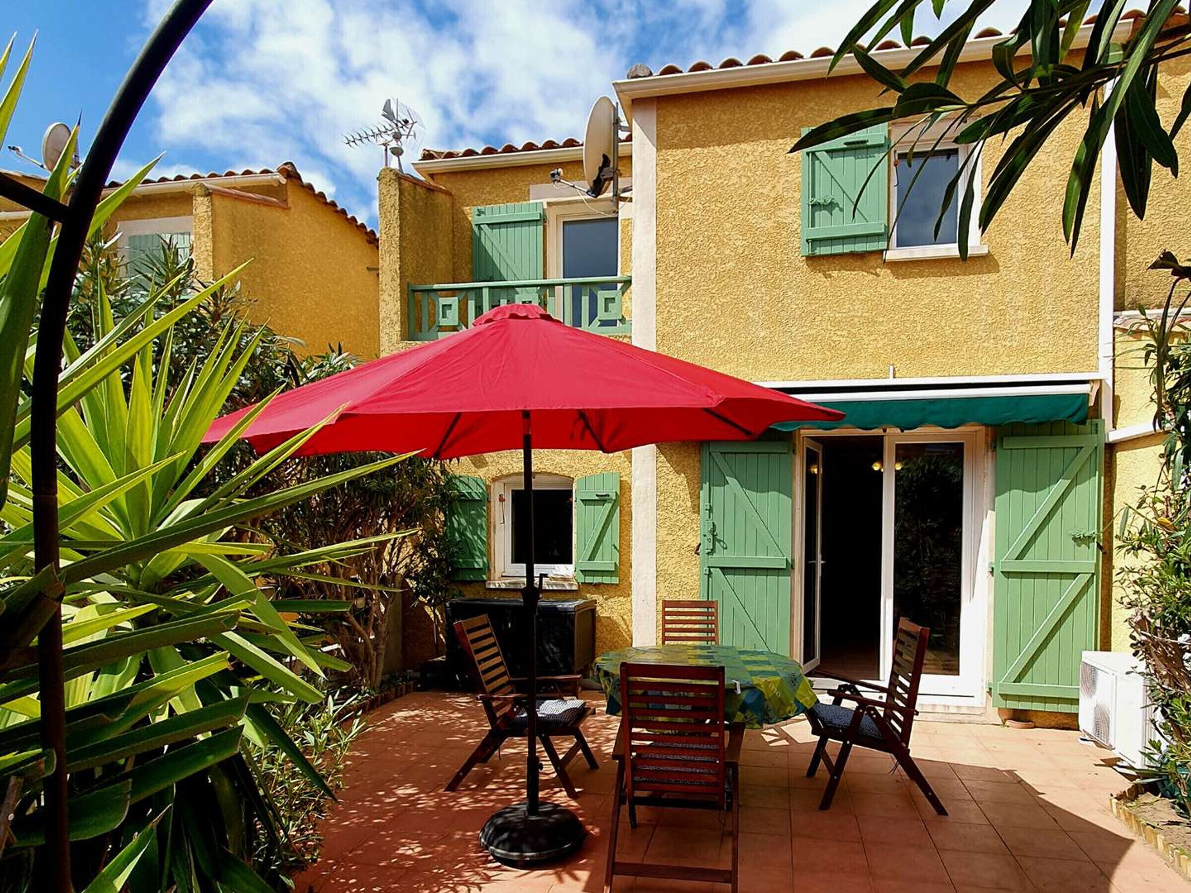 A patio with a red umbrella, outdoor furniture, and a building with green shutters.