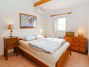 Ein Schlafzimmer mit einem hölzernen Bett, einem Nachttisch mit Lampe, einer Kommode, einem Fenster mit Vorhängen und einem Bild an der Wand.