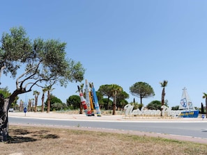 A coastal road with palm trees, a sailboat, and a sign reading "Nautique Plage". - Cozy 3-Bedroom Villa with A/C, Terrace, and BBQ – 350m from Beach in Narbonne-Plage (Narbonne-Narbonne Plage-Narbonne Plage)