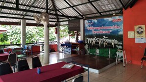 A covered outdoor seating area with tables and chairs, a stage with a backdrop, and a red wall with a calendar and a framed picture. - Phetsangsom Resort (Klaeng)