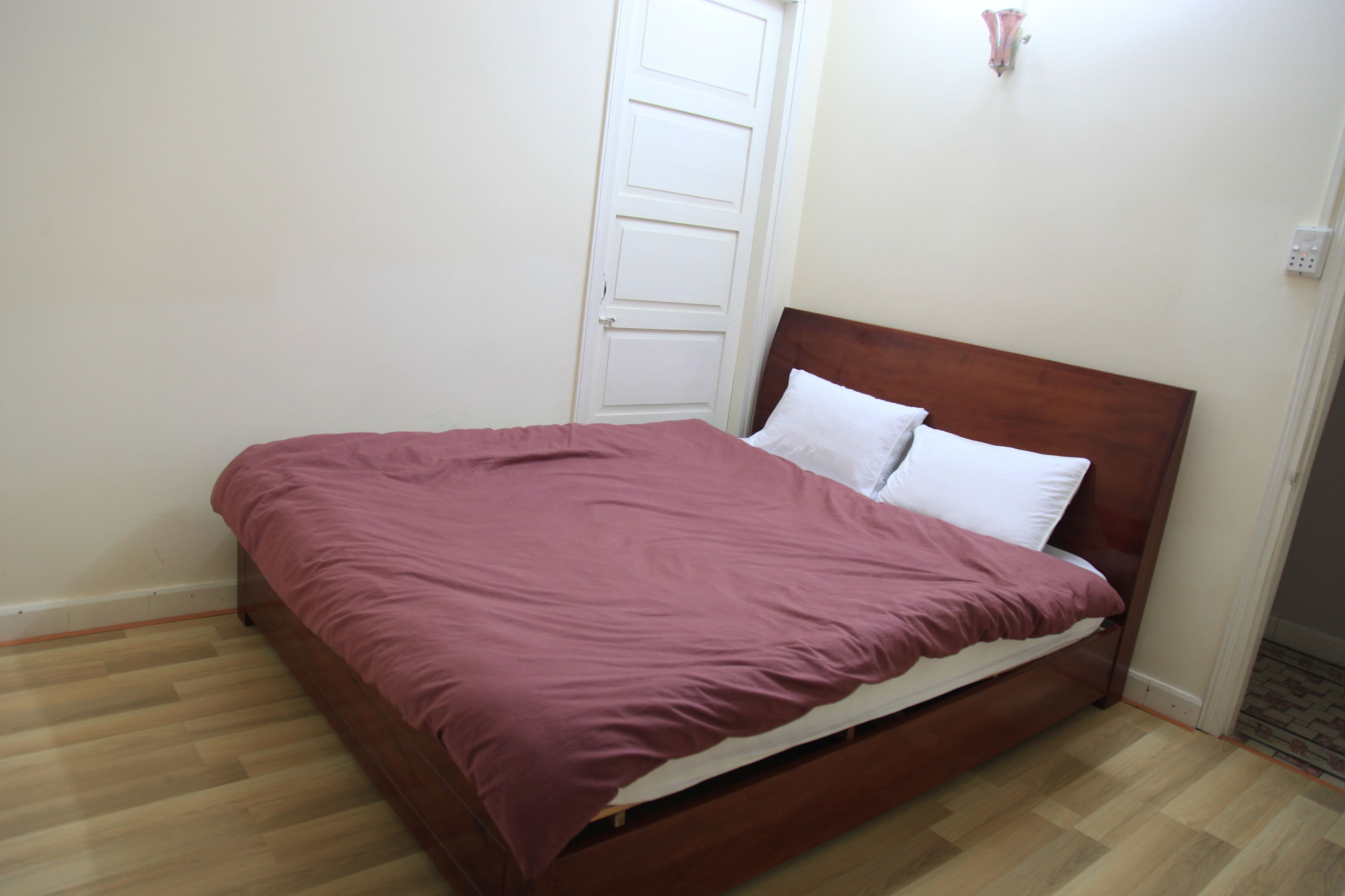 A single bed with a wooden headboard and a pink bedspread in a room with light-colored walls and wooden flooring.