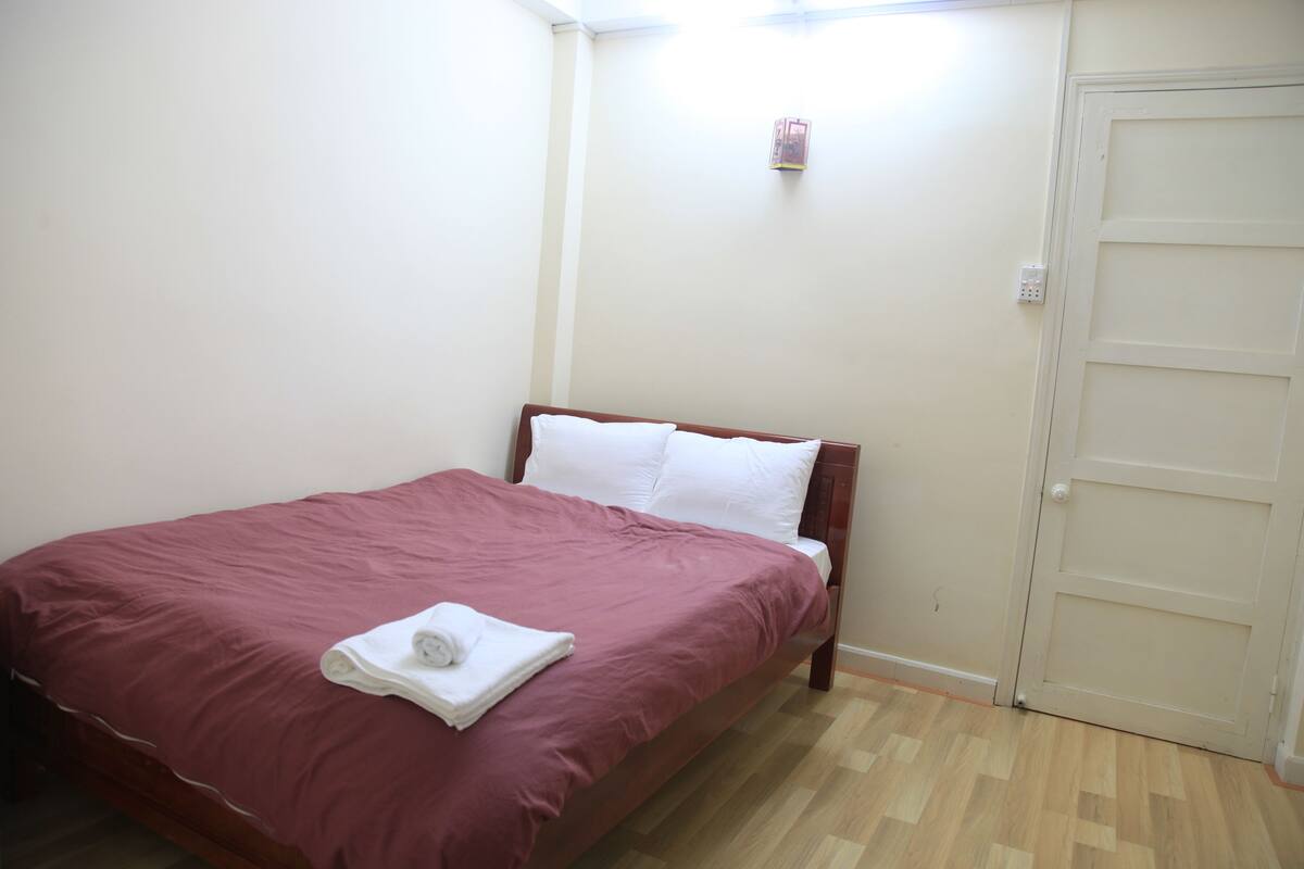 A single bed with a wooden frame and a red bedspread, a white pillow, and a rolled white towel on the bed.