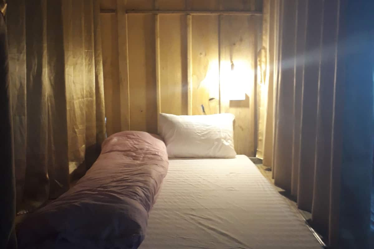 A single bed with a white mattress and a pink blanket, a white pillow, and a bedside lamp.