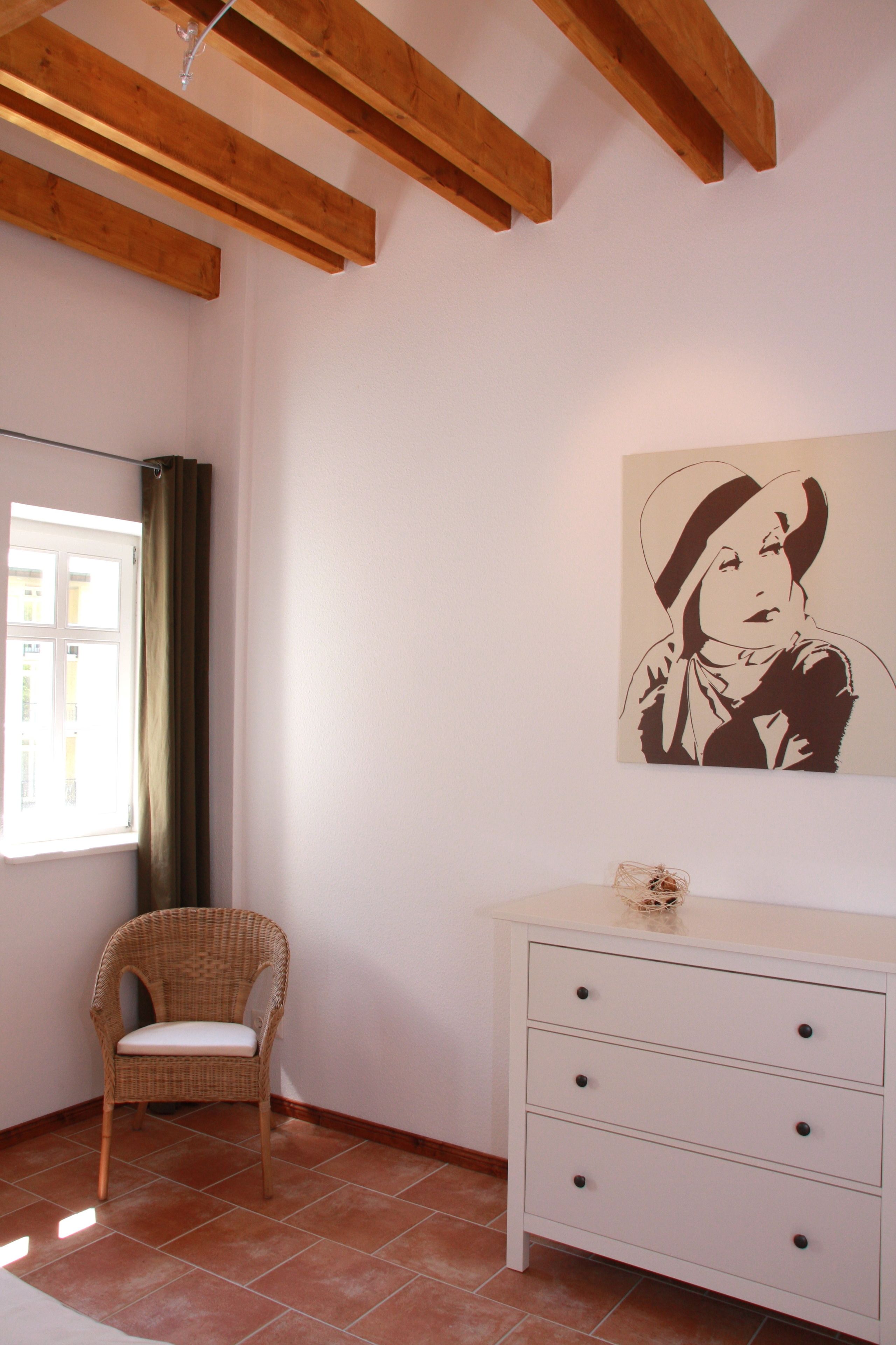 A room with a wicker chair, a white dresser, and a wall-mounted artwork.