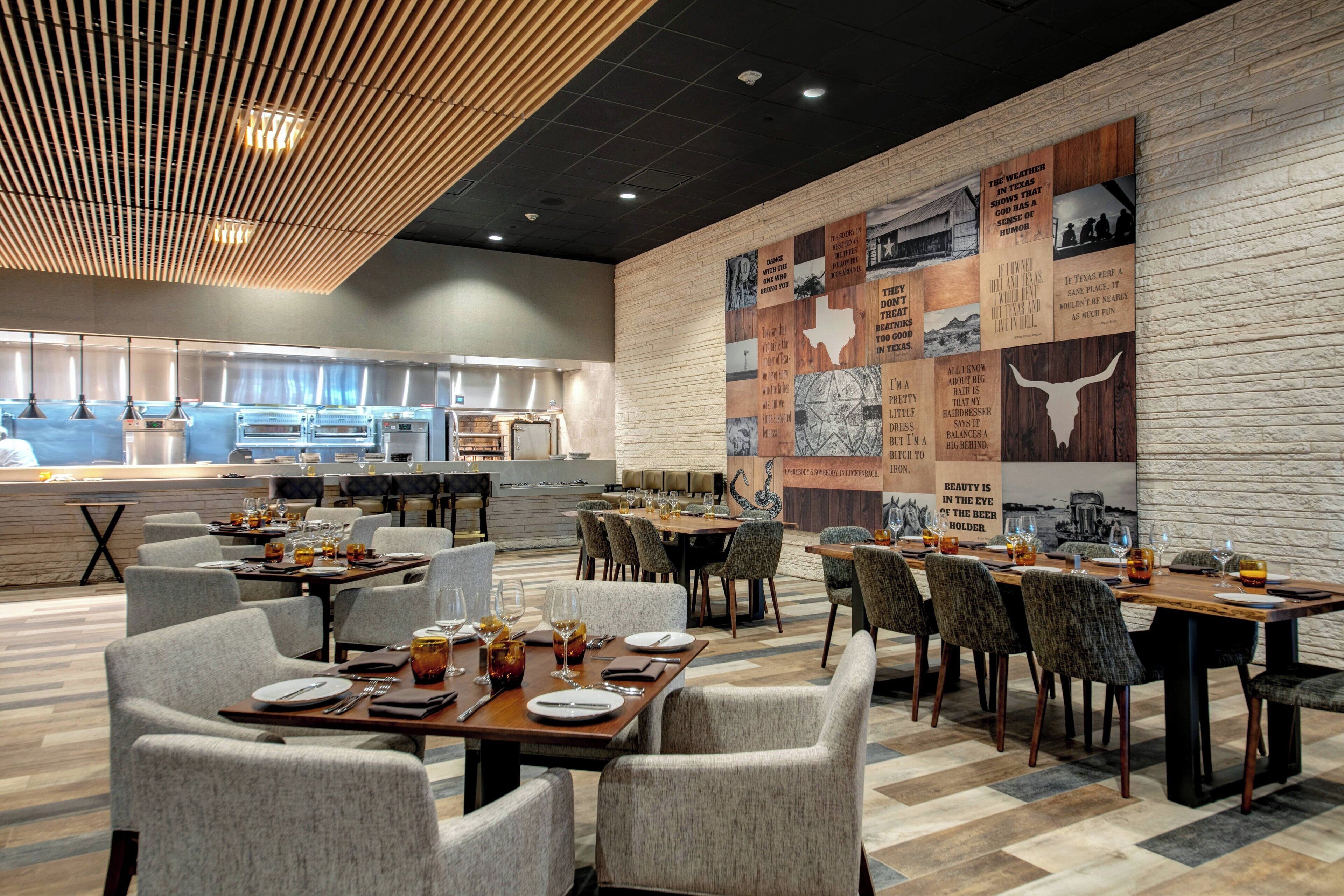 A restaurant with multiple dining tables and chairs, a kitchen area in the background, and a wall with various images and text.