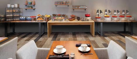 A hotel dining area with a long wooden table set for four, surrounded by grey upholstered chairs.