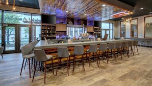 Een bar met houten plafond, barstoelen en flessen op planken.