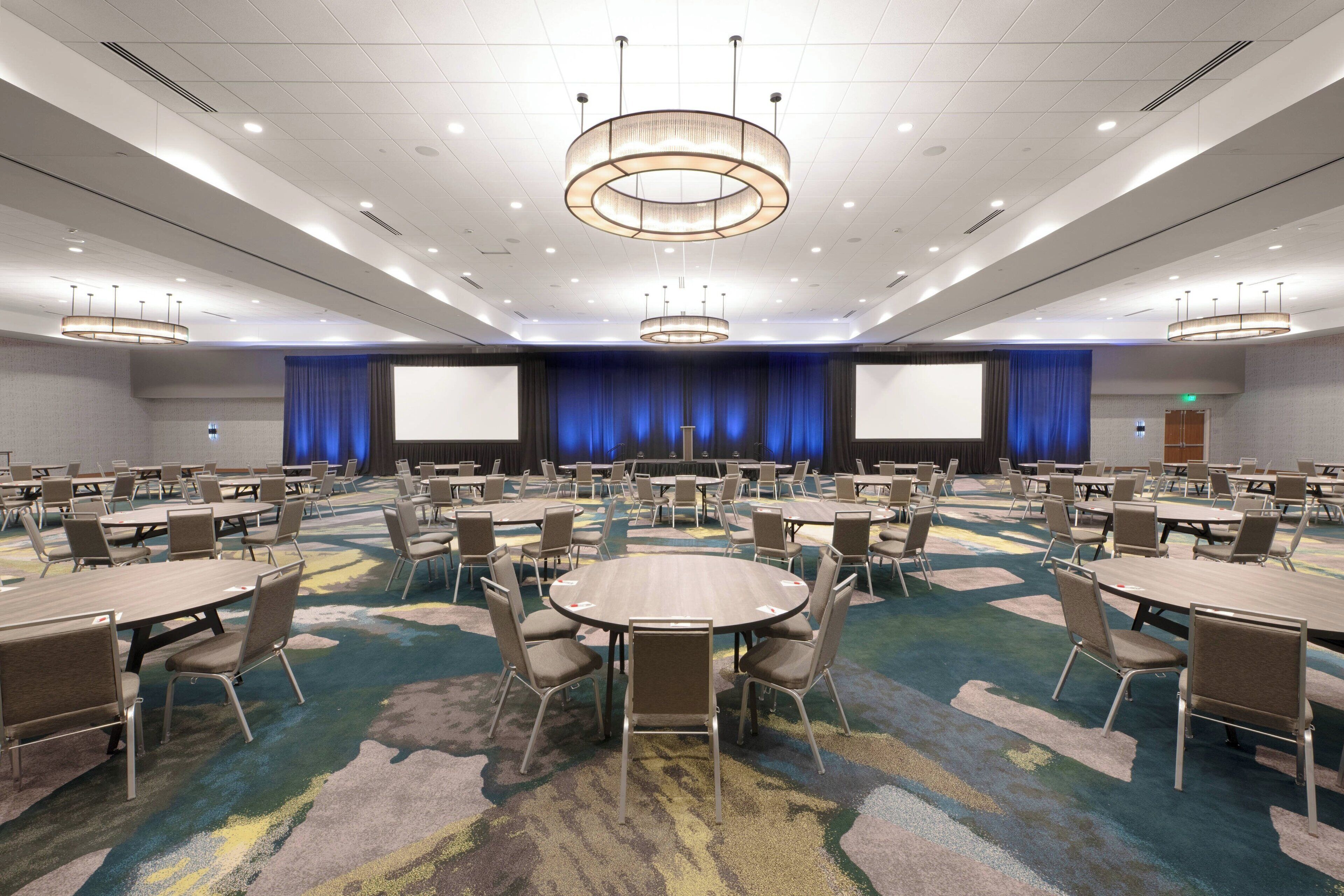 A large conference room with multiple tables and chairs, a stage with a blue curtain backdrop, and a large screen.