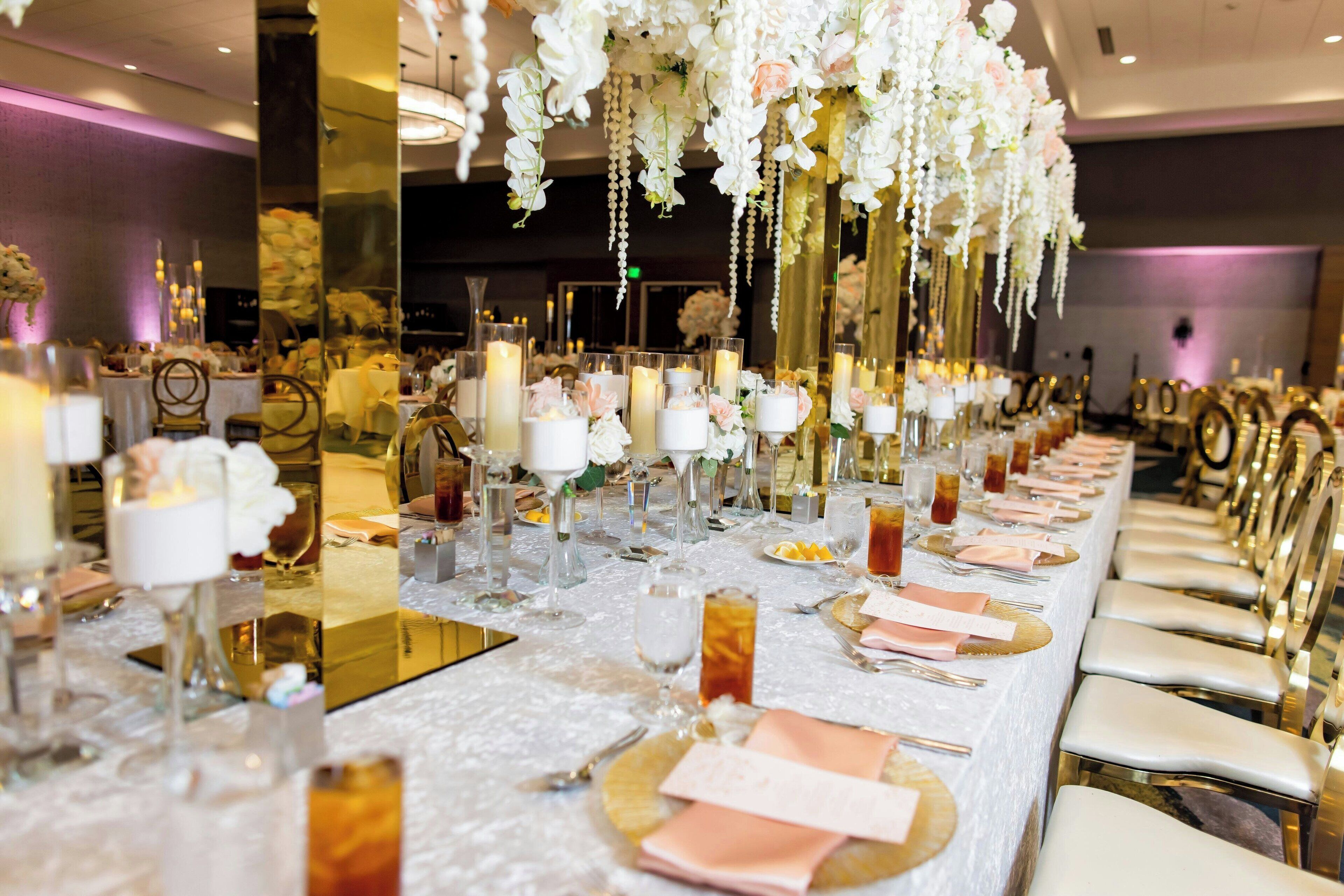 Una larga mesa de banquete con manteles blancos, platos bajos dorados y servilletas rosas.