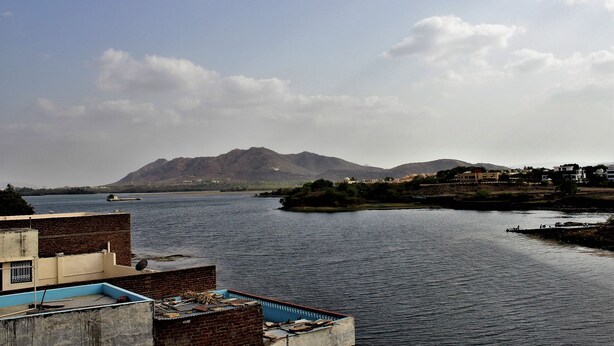 A riverside view with a mountain in the background, a bridge, and buildings along the riverbank. - Rising Shemesh (Udaipur)