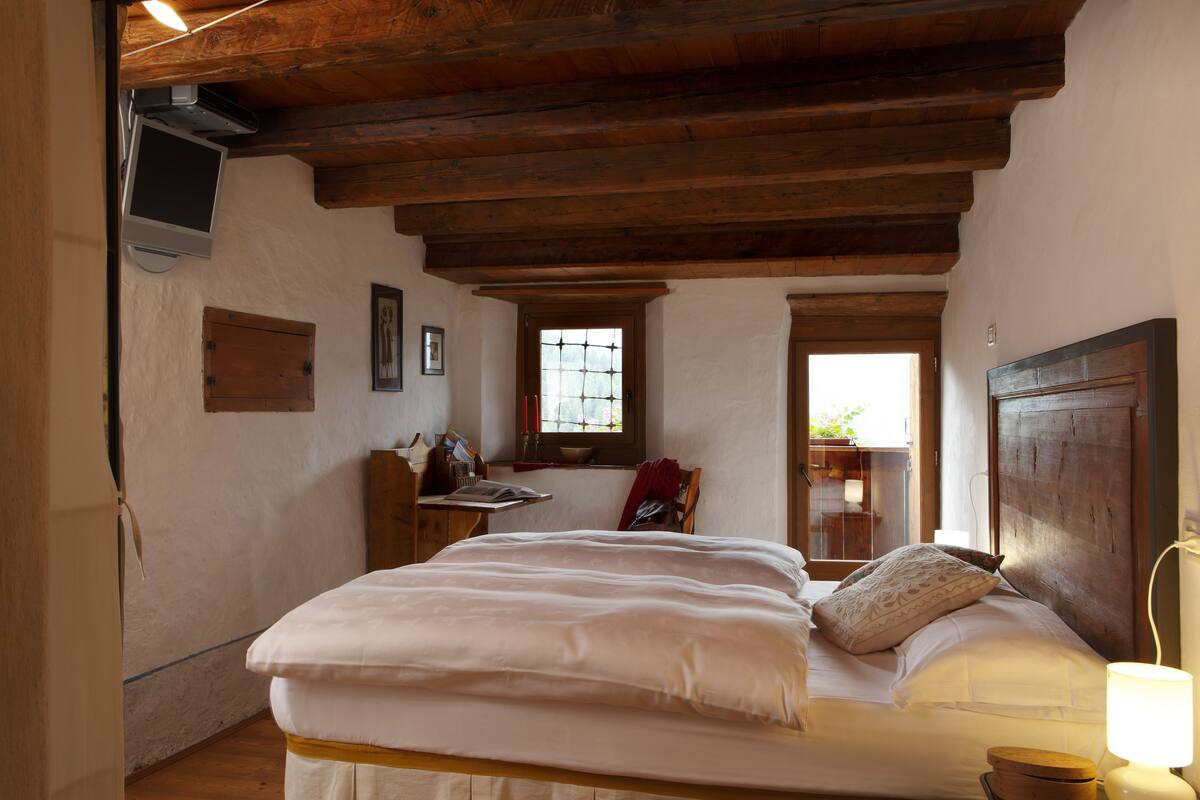 A bedroom with a wooden ceiling, a bed with white linens, a window with a view, and a wooden nightstand with a lamp.
