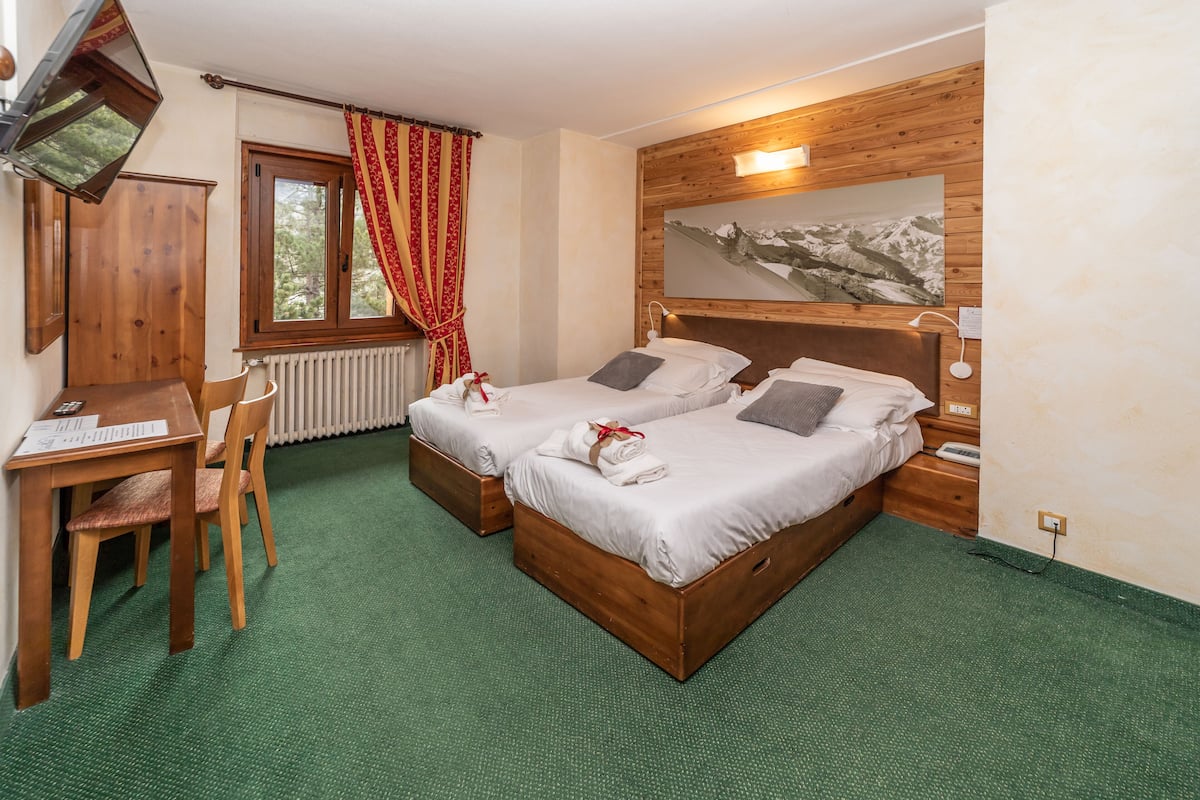 Habitación de hotel con dos camas de madera, un escritorio con silla, un radiador, una ventana con cortinas y una pared de madera con un cuadro.