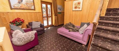 A wooden interior with a staircase, two sofas, a patterned carpet, and a door leading outside.