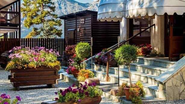 A courtyard with wooden benches, potted plants, and a staircase leading to a building with a dome structure. - Hotel Gran Baita (Sauze d'Oulx)