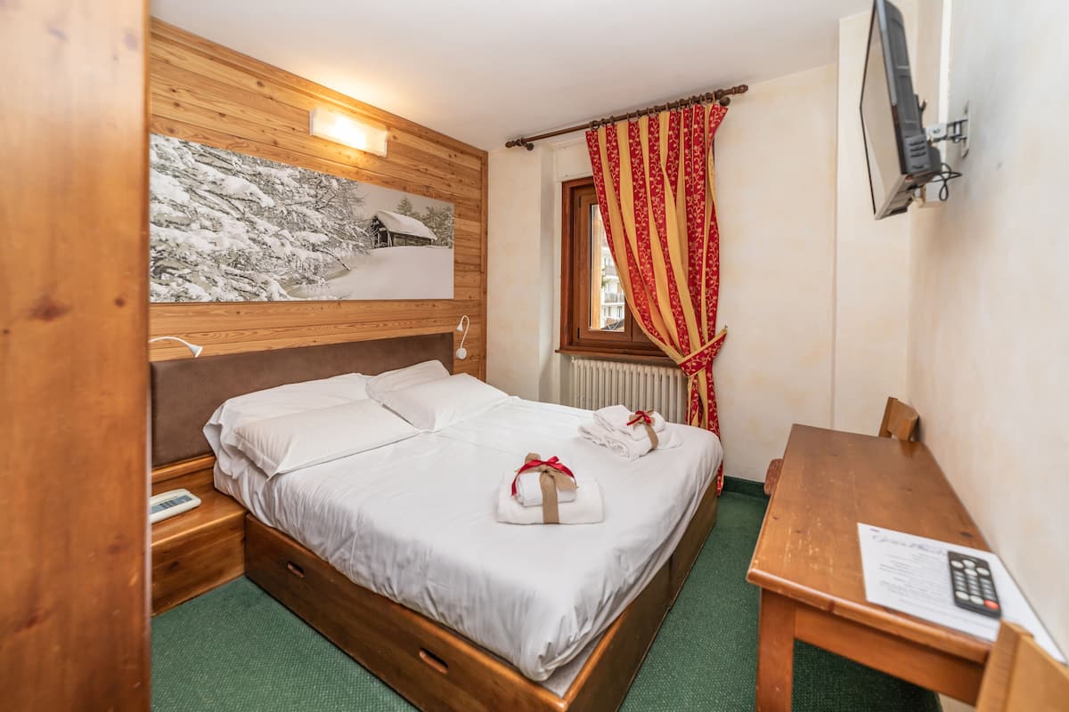 Habitación de hotel con una cama de madera, una mesita de noche pequeña, un escritorio con una silla, una ventana con cortinas y un televisor montado en la pared.