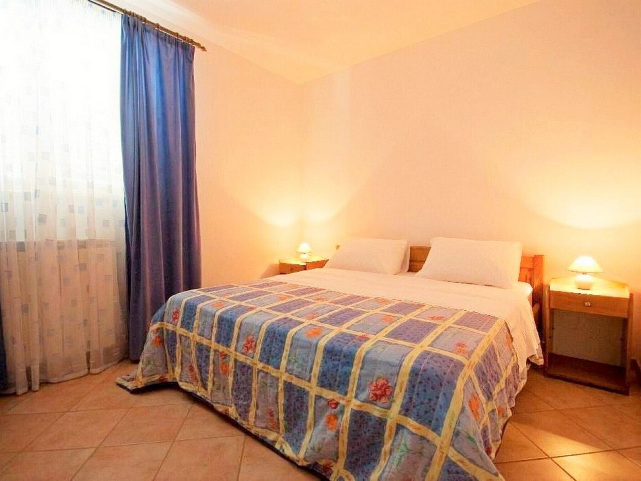 Une chambre à coucher avec un lit, deux tables de chevet avec des lampes, une fenêtre avec des rideaux et un plancher carrelé.