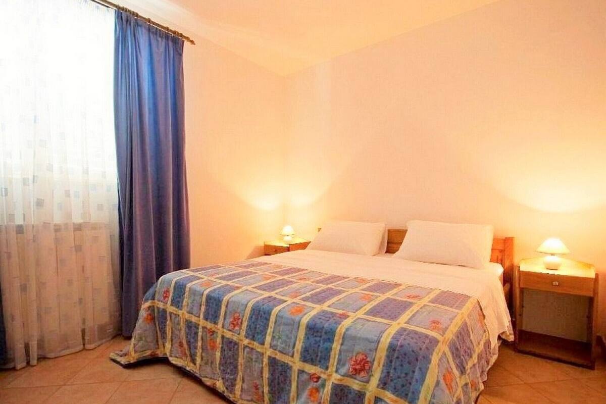 A bedroom with a bed, two bedside tables with lamps, a window with curtains, and a tiled floor.