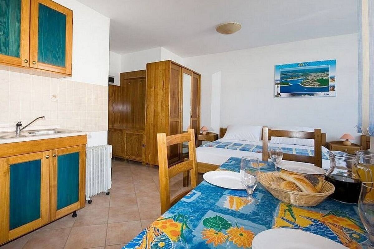 A room with a bed, a dining table with a tablecloth, a kitchen area, and a view of the sea.