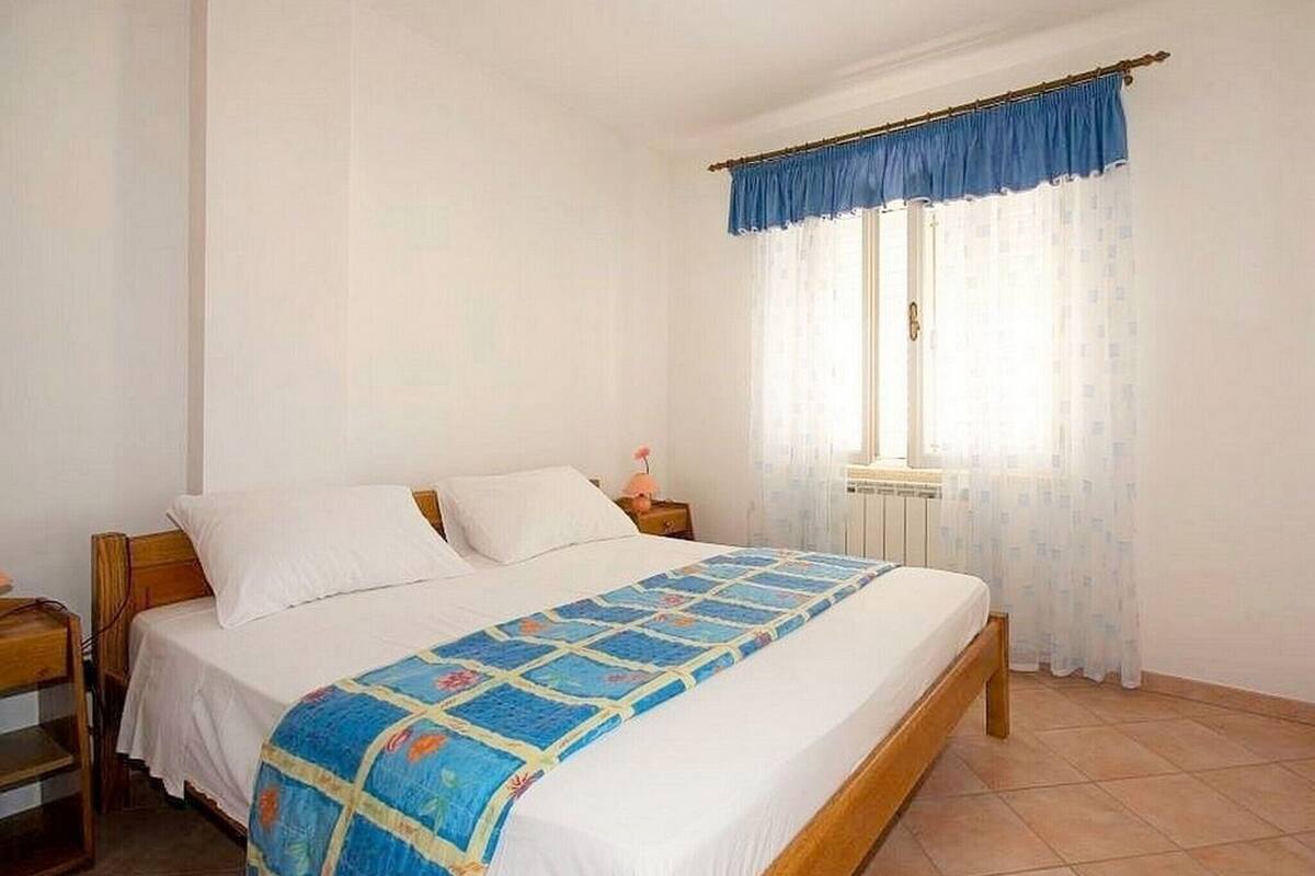 A bedroom with a bed, a nightstand, a window with curtains, and a radiator.