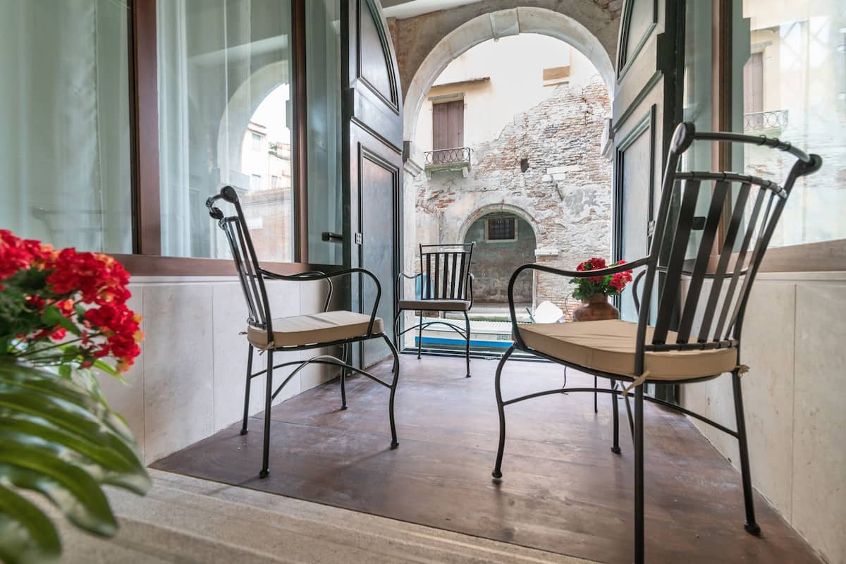 A balcony with two chairs and a table, overlooking a courtyard with a stone wall and a door.