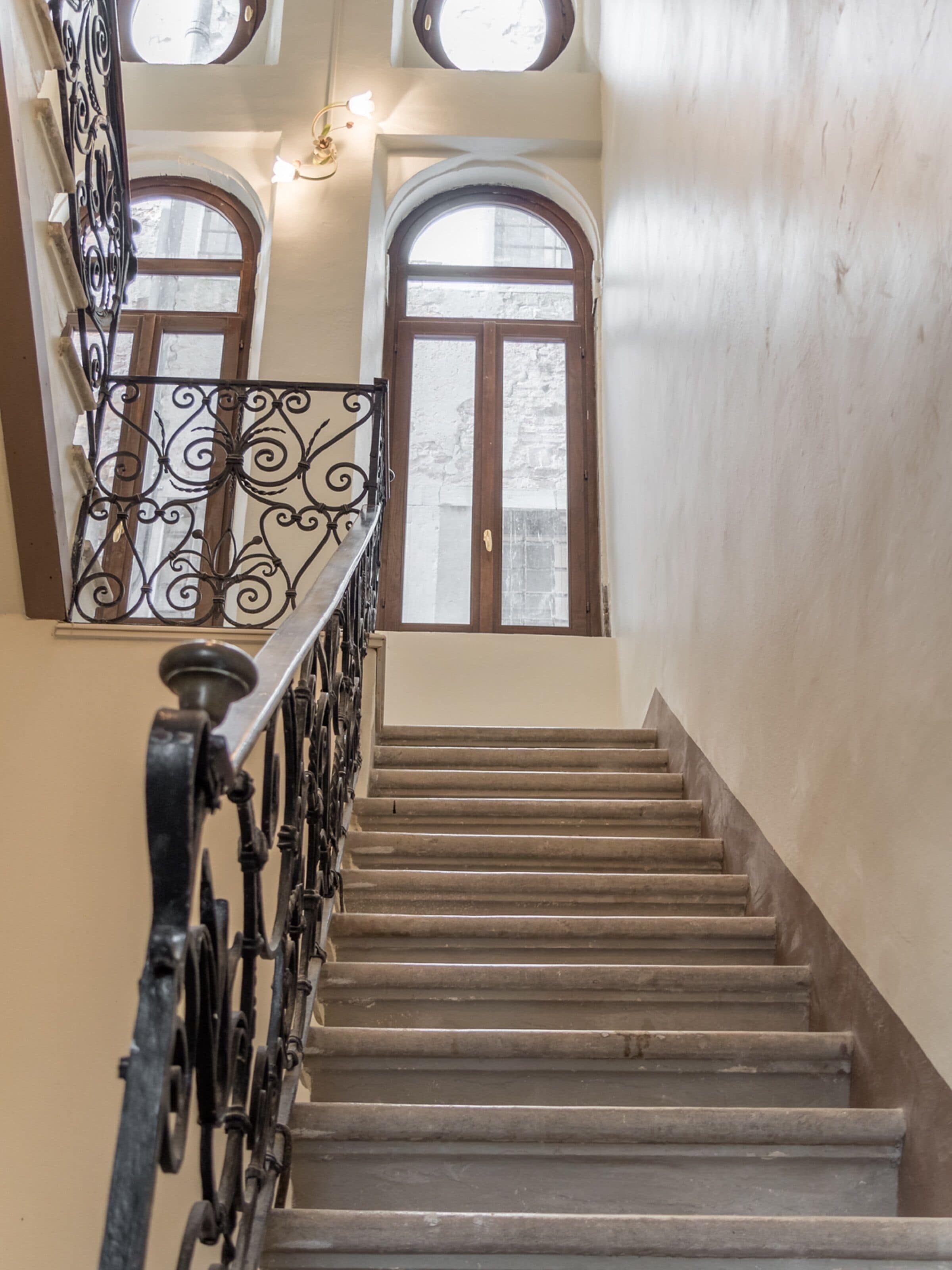 A staircase with a wrought iron railing, wooden steps, and a view of two arched windows with wooden frames.