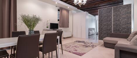 Una cocina moderna con armarios blancos, un comedor con mesa y sillas, y una sala de estar con sofá y alfombra estampada.