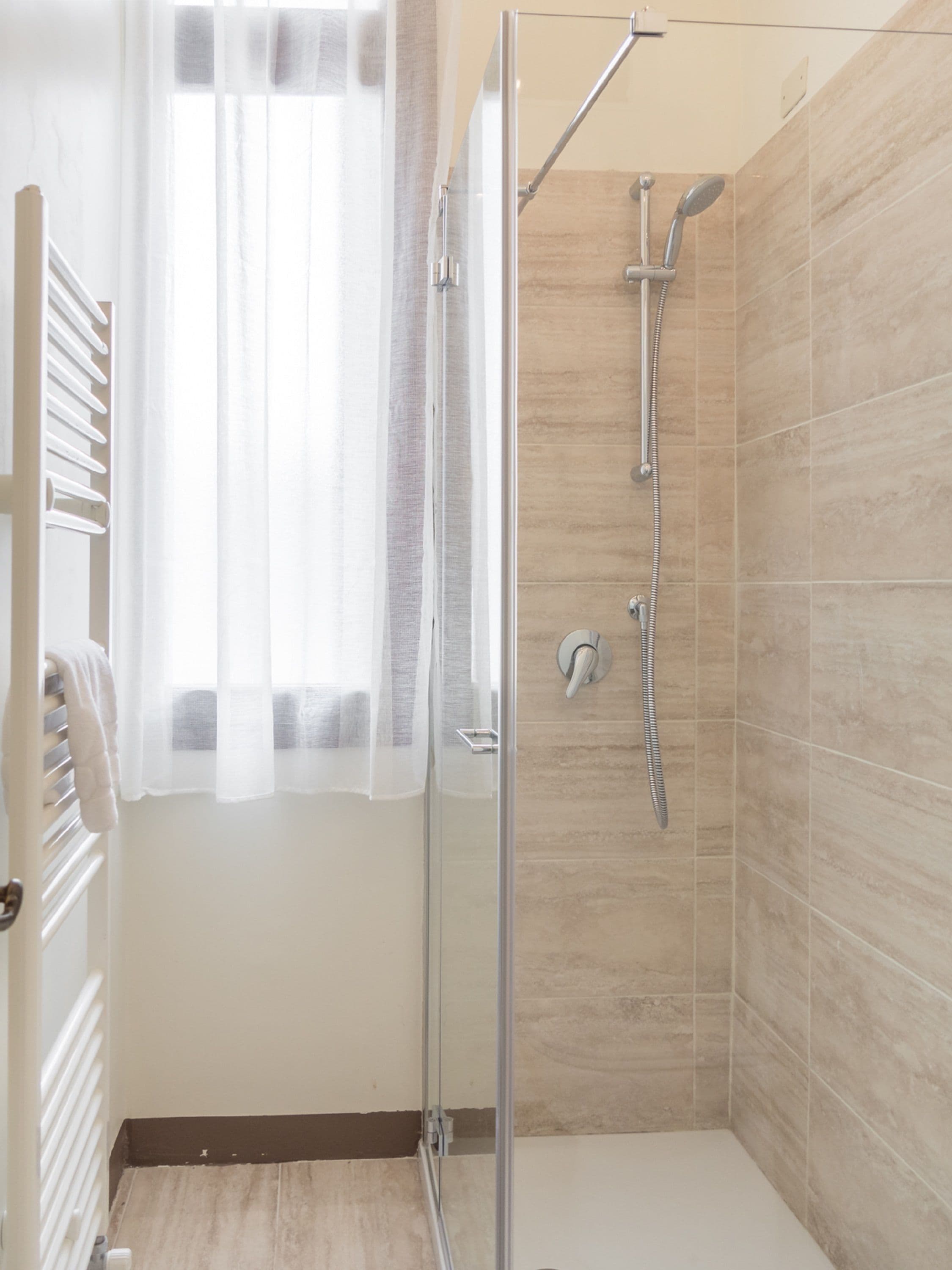 A modern bathroom with a glass shower enclosure, a towel rack, and a window with sheer curtains.