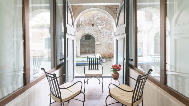 A corridor with two chairs and a table, leading to a courtyard with a brick wall and a window. - Riva Palace Apartments by Wonderful Italy (Venice)