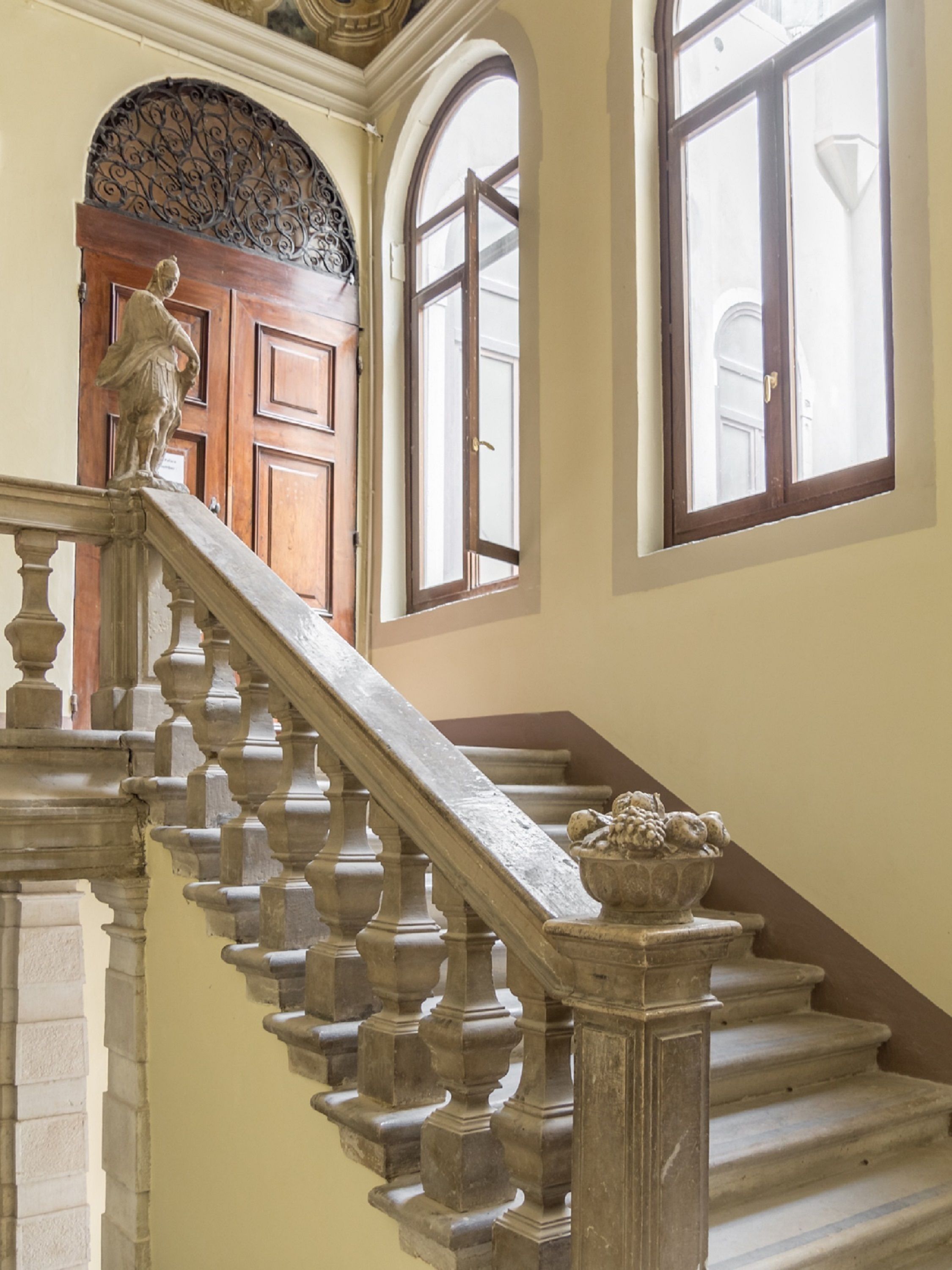 Un magnifique escalier, orné de balustres travaillés et surmonté d’une balustrade en pierre, mène à une porte en bois surmontée d’un arc décoratif.