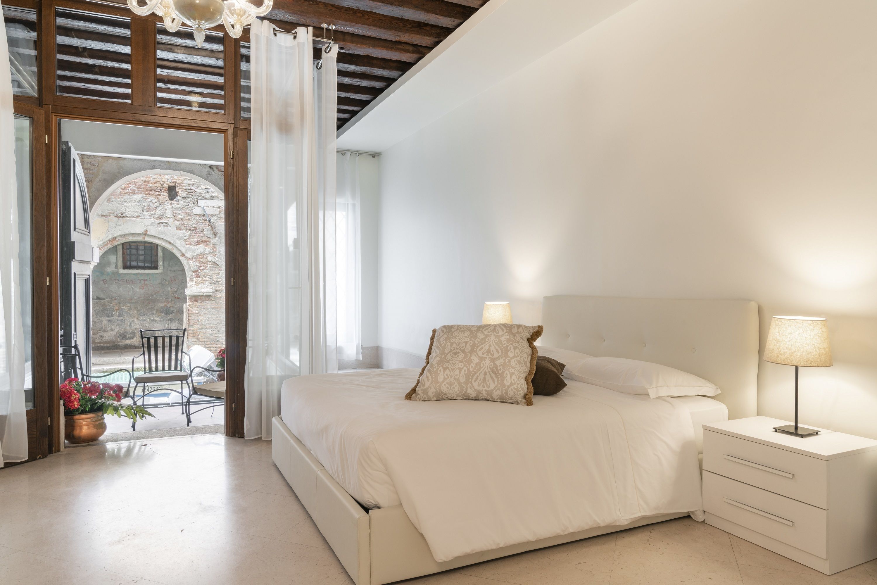 Un dormitorio con cama, mesita de noche y lámpara. Se ve un área exterior con mesa y sillas.