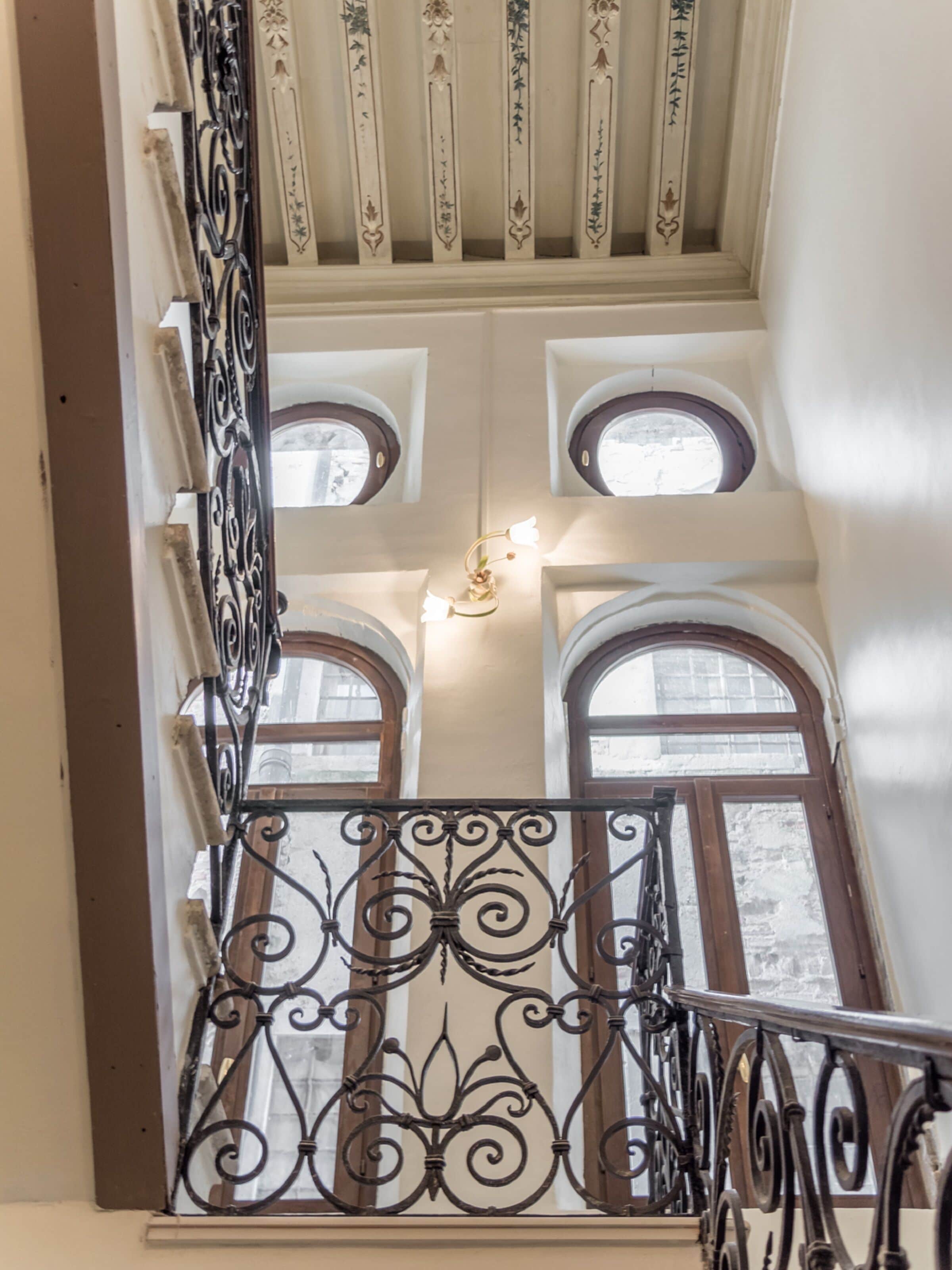 Eine Treppe mit aufwendiger schwarzer Eisenbalustrade und mehreren Fenstern mit Rundbögen.