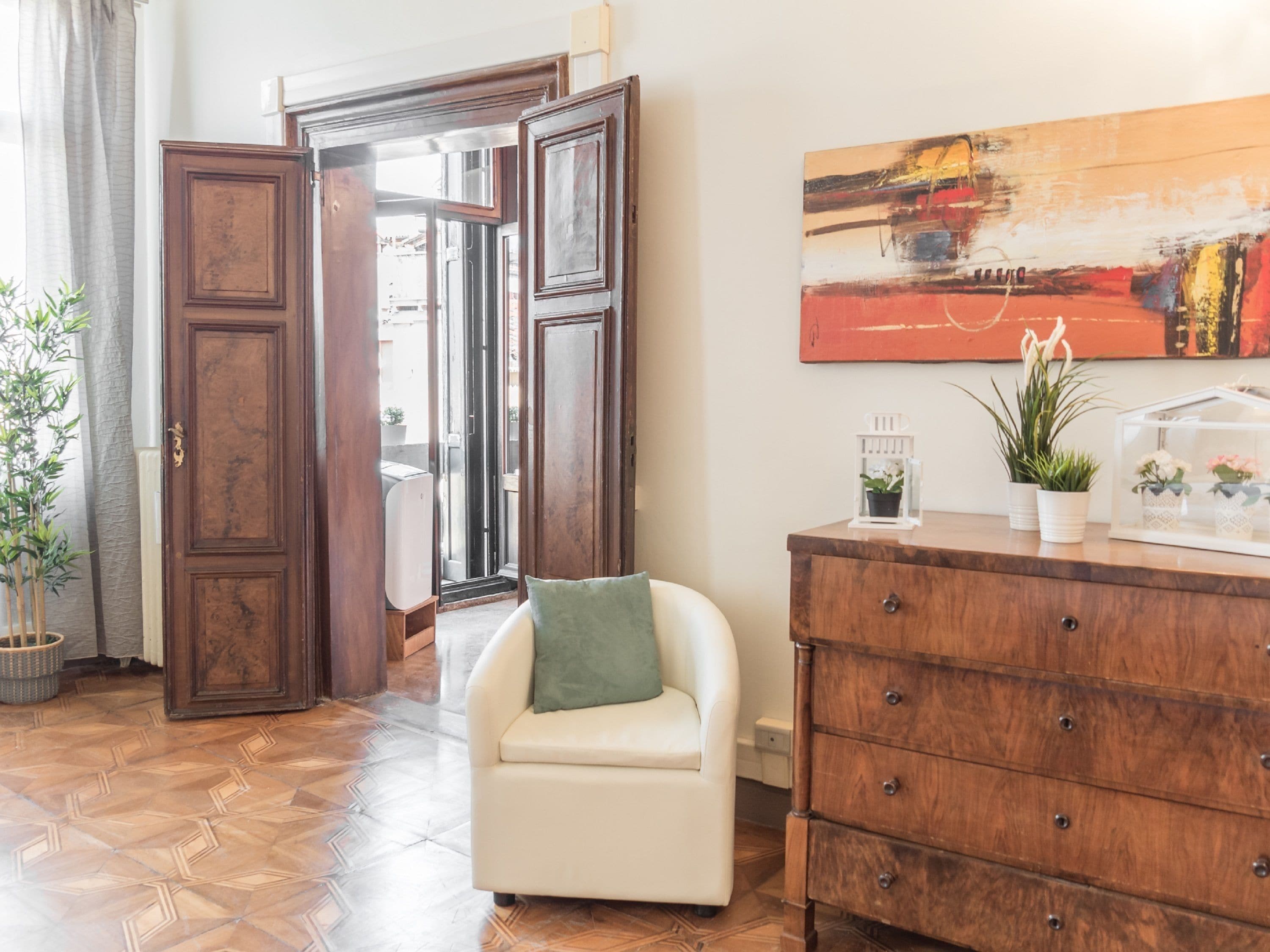 A room with a wooden floor, a white armchair, a wooden dresser, and a painting on the wall.