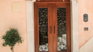 A wooden double door with a decorative arch above it, a small potted plant to the left, and a metal doormat in front. - Casa Turchetti - Taormina (Taormina)