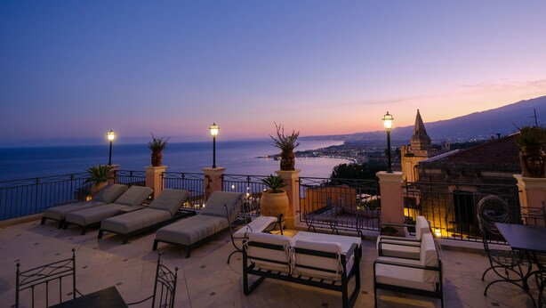 A terrace with lounge chairs and tables, overlooking a cityscape and the sea at dusk. - Casa Turchetti - Taormina (Taormina)